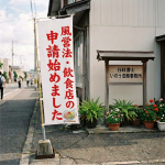 Banner reading 「風営法の申請始めました」 and 「許可申請・ご相談」 outside a sign reading 「行政書士 佐藤事務所」